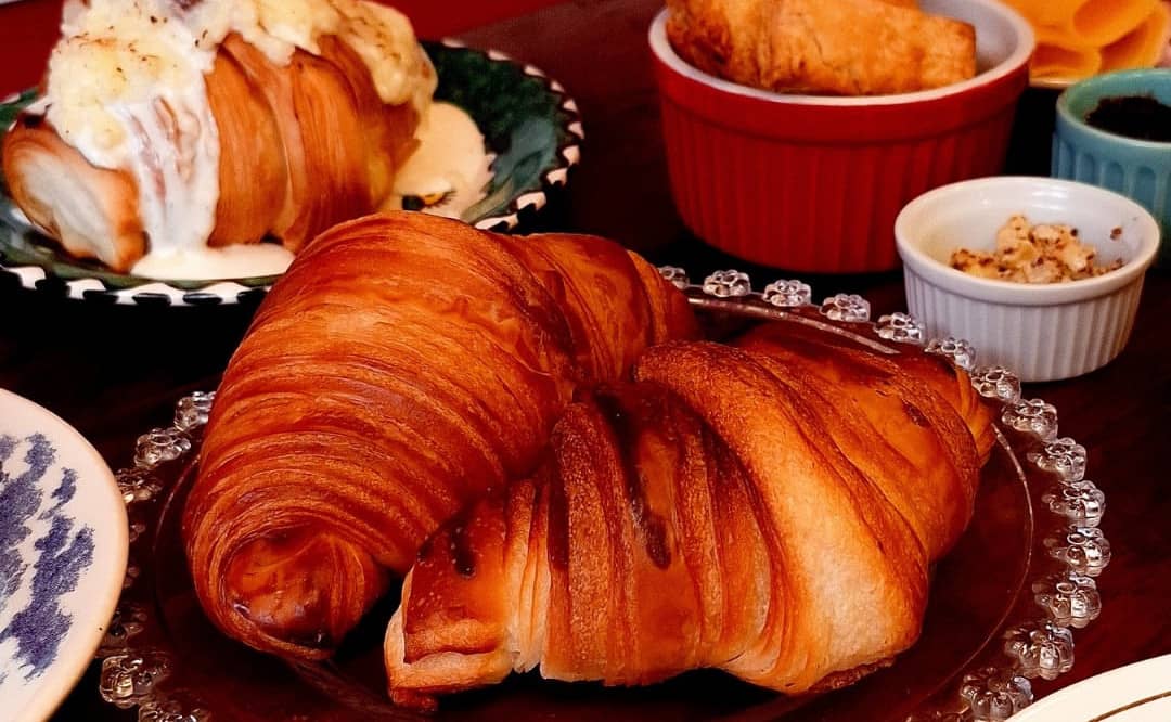 Dois mini croissants num prato de vidro de borda trabalhada; ao fundo, potinhos, queijo e presunto na mesa.