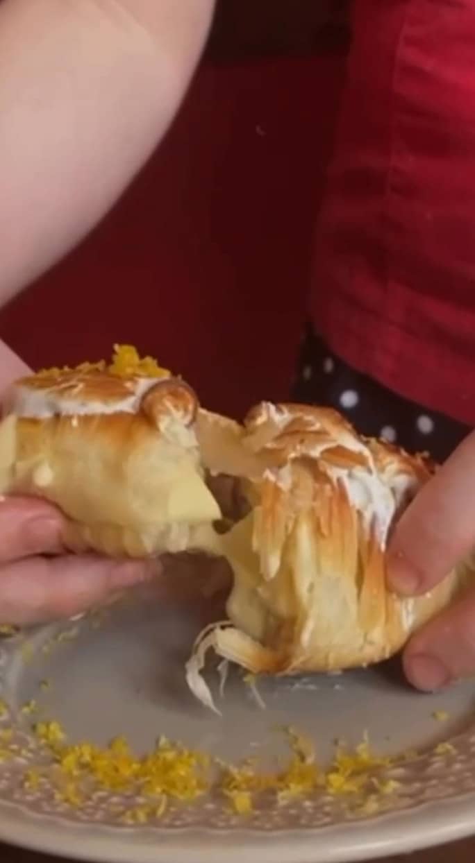 Duas mãos abrindo um croissant recheado de creme, com raspas de limão no prato.