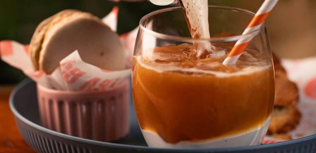 Café gelado sendo servido num copo com merengue no fundo, ao lado de um macaron no papel listrado da casa.