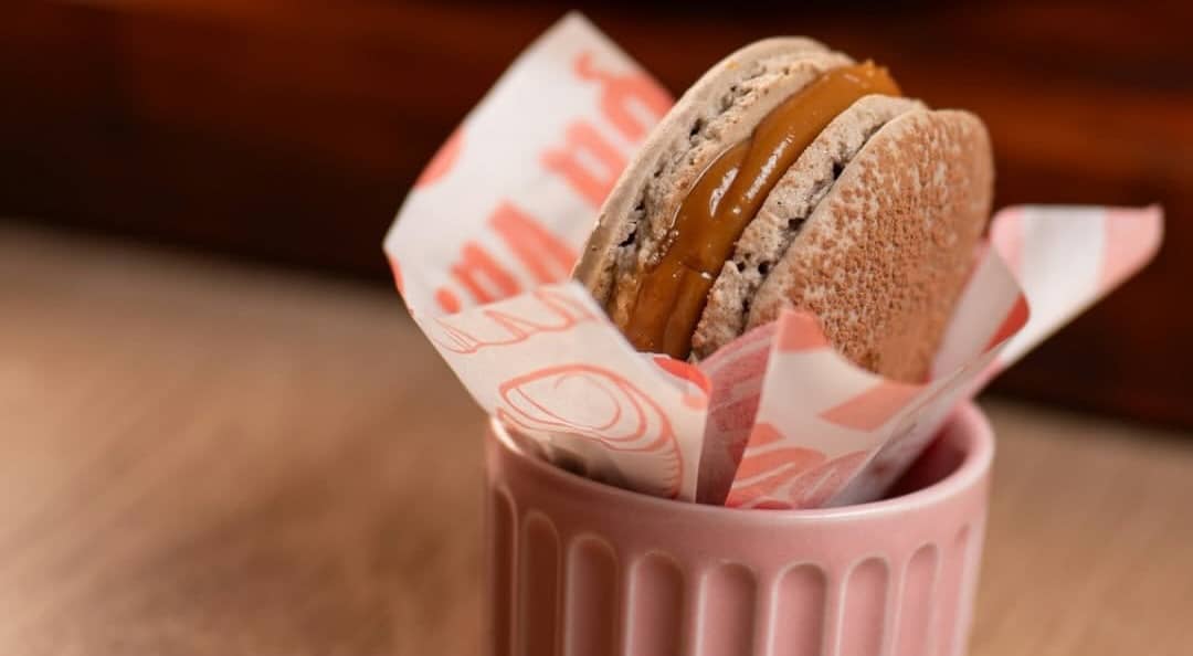 Macaron recheado com doce de leite, de pé num potinho rosa forrado com o papel da casa.