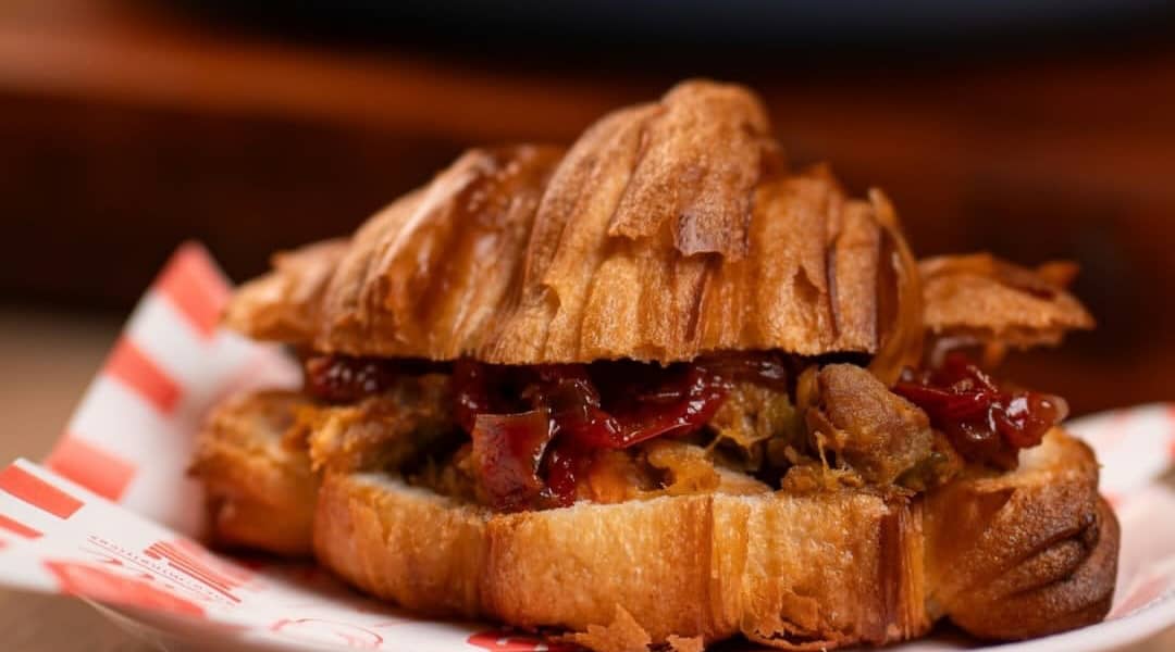 Croissant aberto recheado com carne de porco desfiada e chutney de tomate-cereja, num pratinho com papel listrado.
