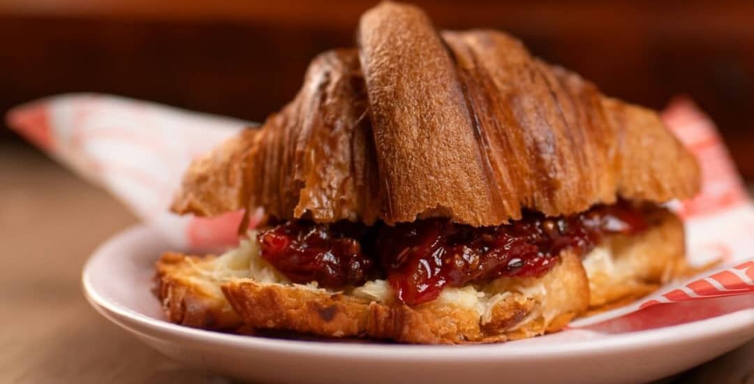 Croissant aberto recheado com queijo curado e chutney de tomate-cereja, num pratinho com papel listrado.